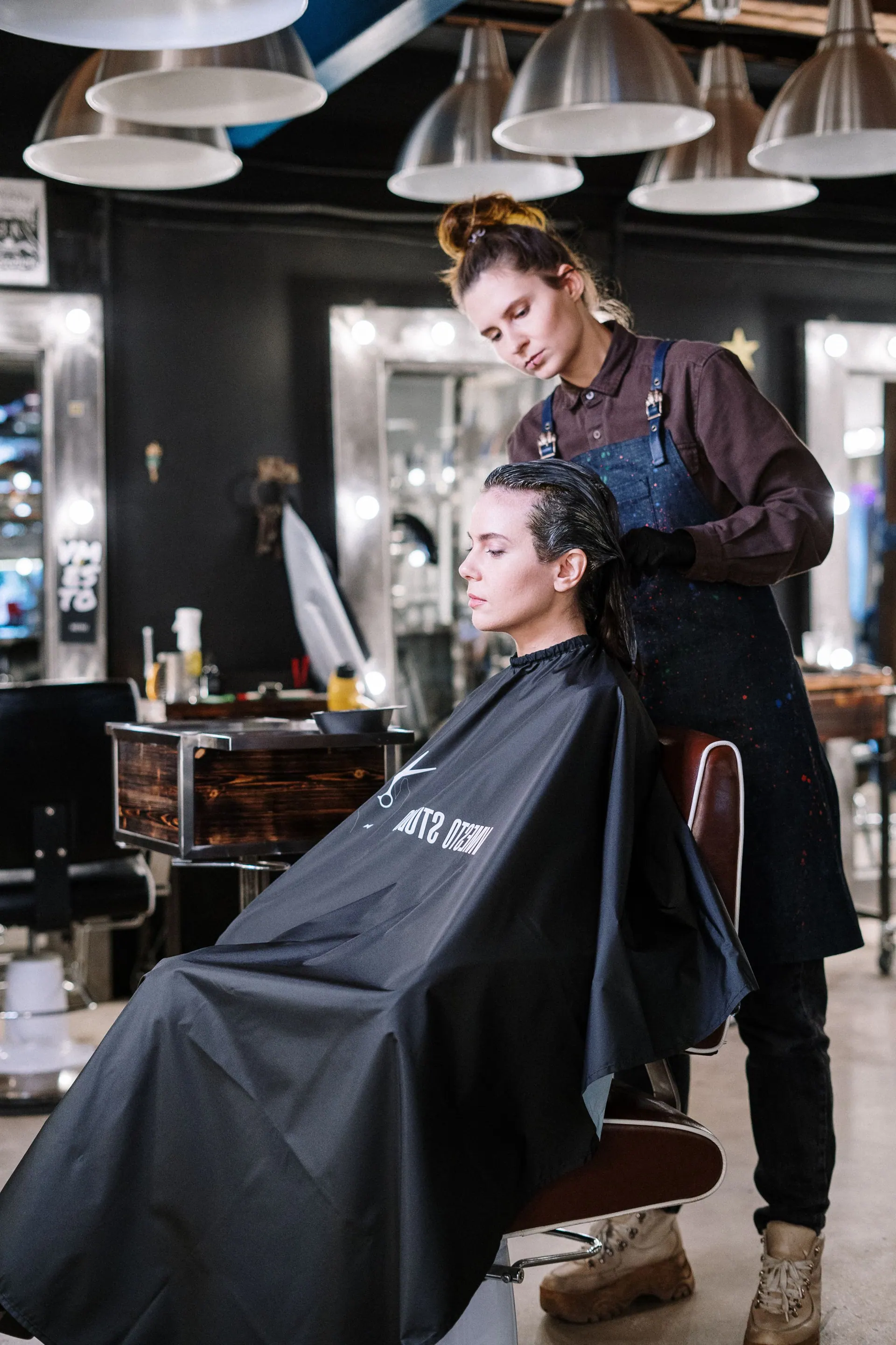 Een kapster in schort knipt een klant in een donkere salon met spiegels en lampen