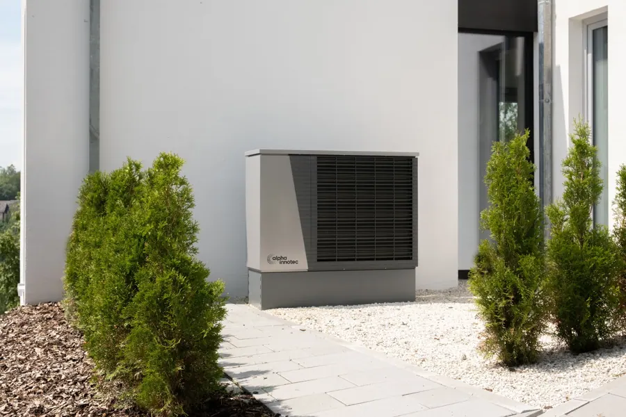 Buitenunit van een warmtepomp tegen een witte gevel tussen coniferen